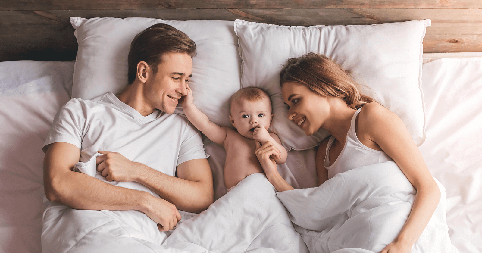 Due adulti e un bambino piccolo su un letto con cuscini bianchi e coperta grigia.