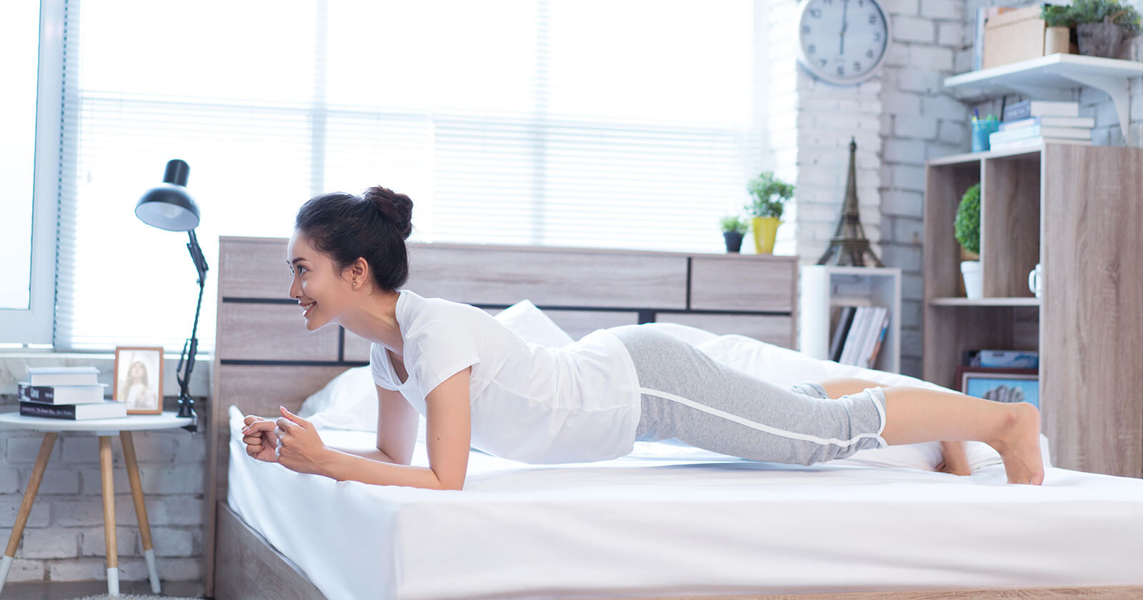 Donna in abbigliamento chiaro appoggiata sugli avambracci e sulle punte dei piedi sopra un letto.